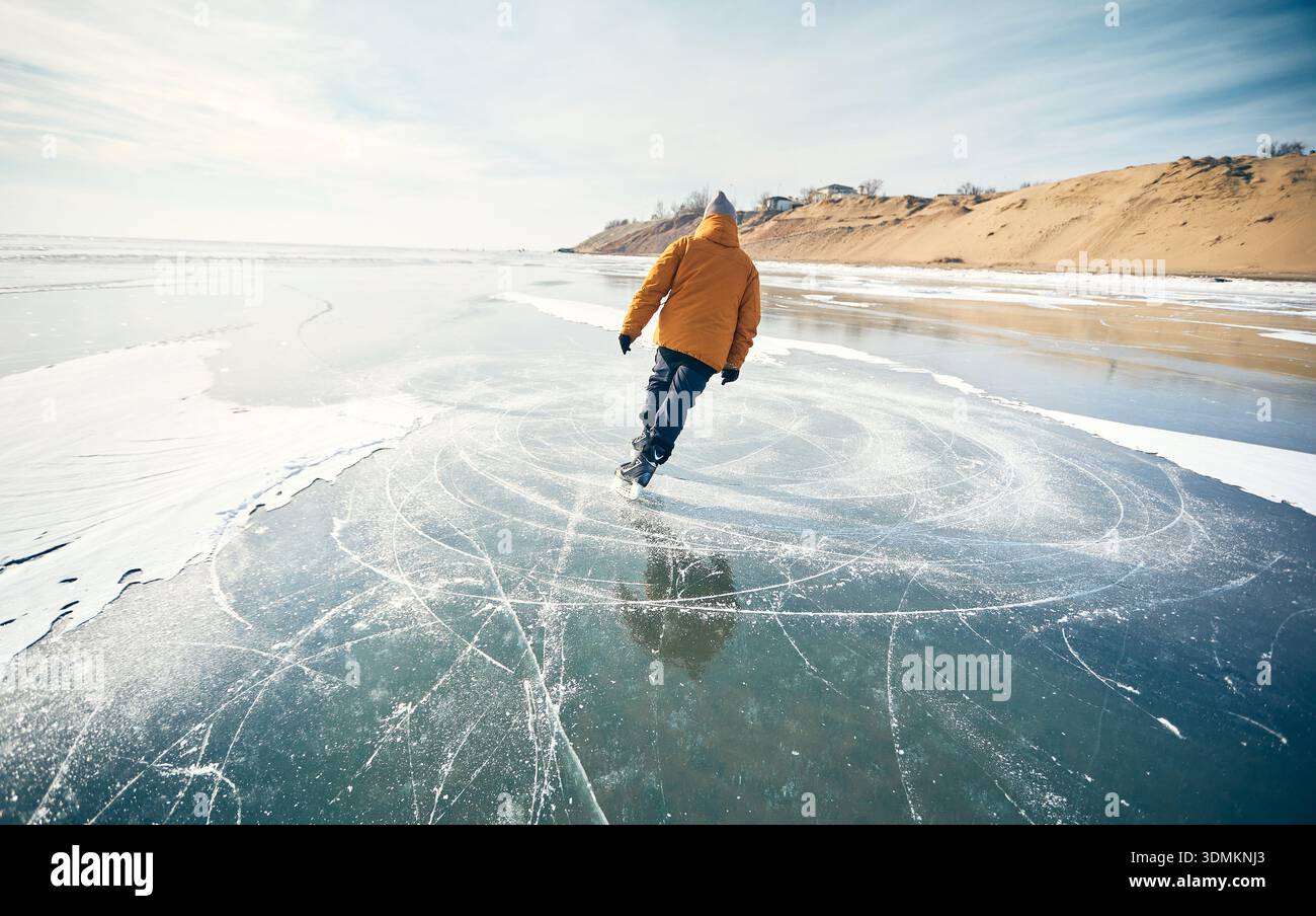 Homme athlète en veste jaune patin sur glace sur le lac gelé avec des fissures et de la neige à l'heure d'hiver Banque D'Images