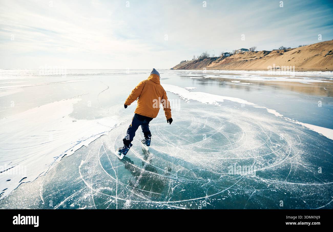 Homme athlète en veste jaune patin sur glace sur le lac gelé avec des fissures et de la neige à l'heure d'hiver Banque D'Images
