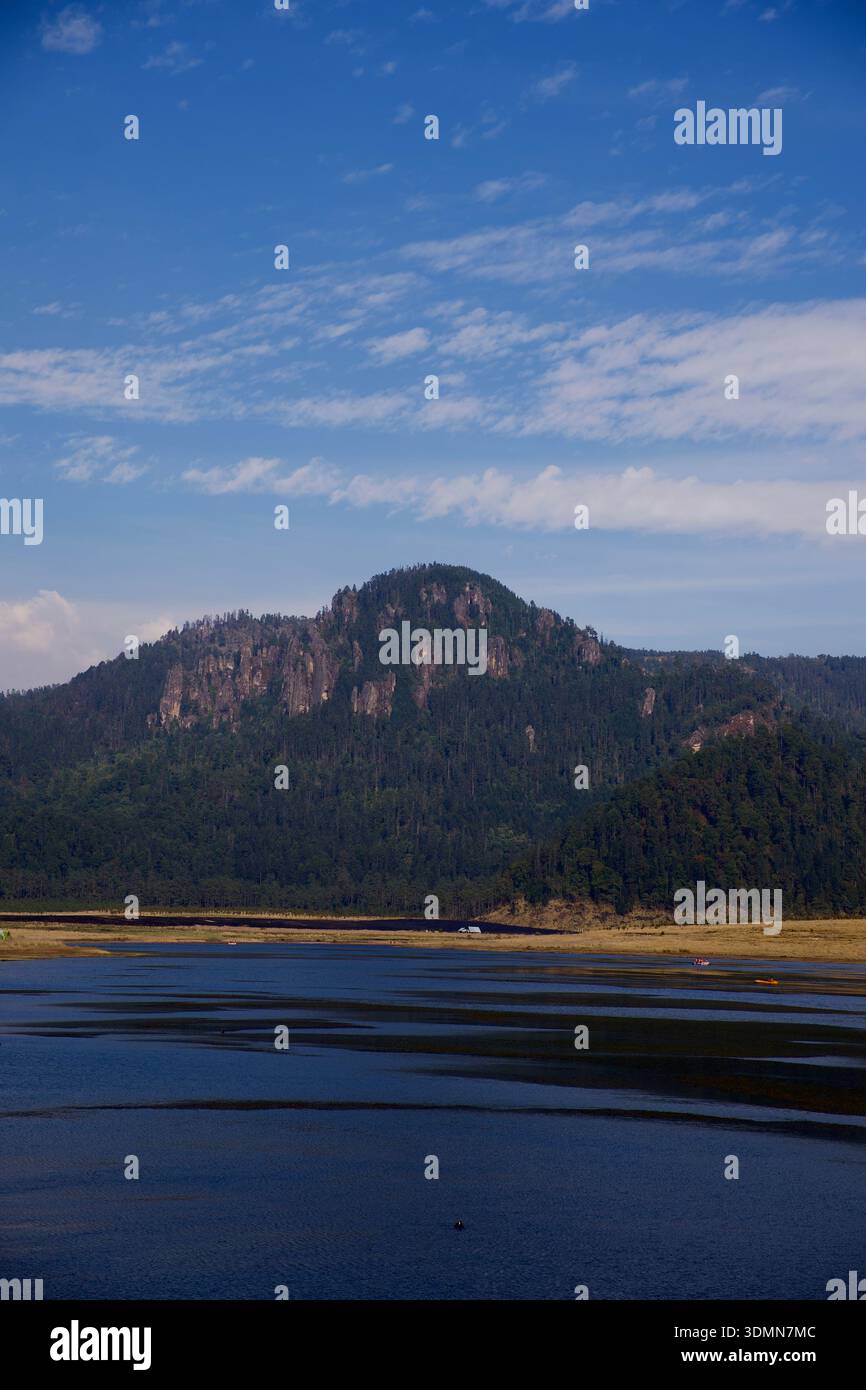 'Montagne boisée s'élevant au-dessus d'un lac calme sous un ciel bleu avec des nuages dispersés, paysage naturel pittoresque. » Banque D'Images