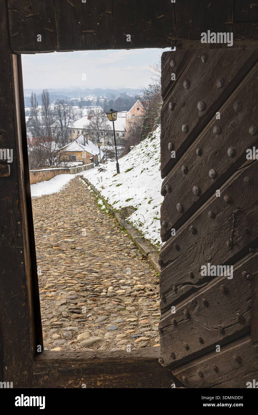 Une scène de village tranquille présente un chemin pavé enneigé vu à travers une grande porte en bois cloutée. Lampadaires bordent le chemin vers des maisons confortables an Banque D'Images