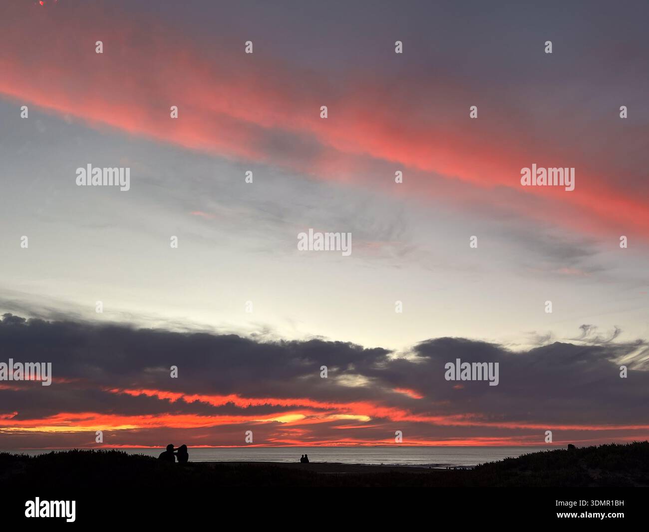 Trois couples s'assoient ou se tiennent debout en silhouette sur le rivage ; au-dessus de l'océan Pacifique, les nuages brillent en rouge sur leurs bords les plus bas - Image de stock capturée avec un smartphone