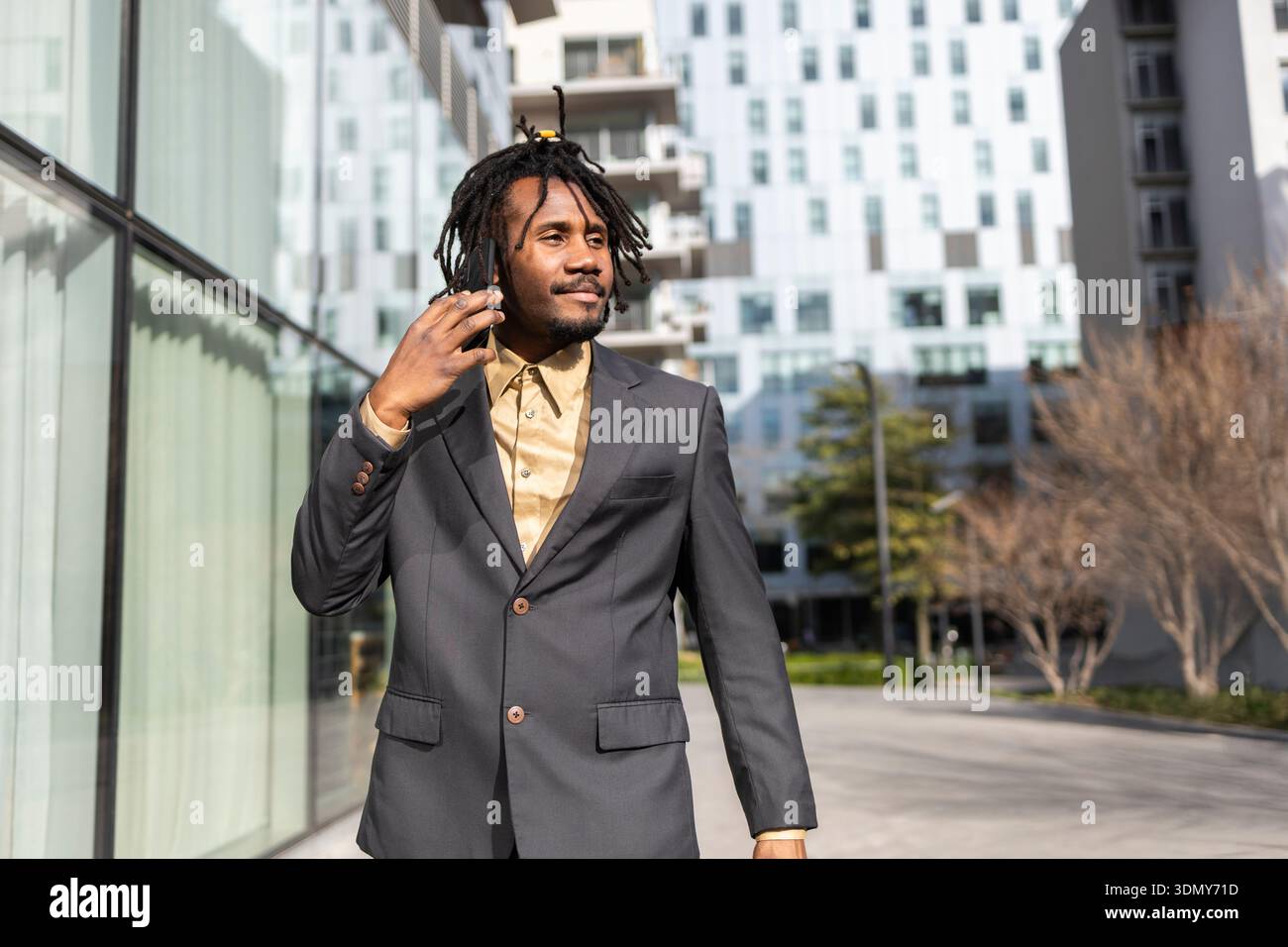 Homme d'affaires afro-américain avec des dreadlocks portant un costume gris et une chemise beige, parlant sur un smartphone. Il marche dehors devant un moderne Banque D'Images