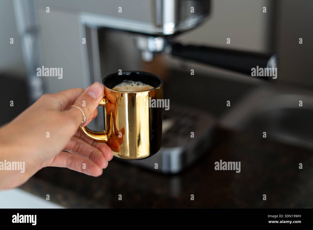 Main de femme ramassant une tasse d'or de café fraîchement infusé à partir d'une machine à café maison argentée - routine matinale élégante, scène de cuisine confortable Banque D'Images