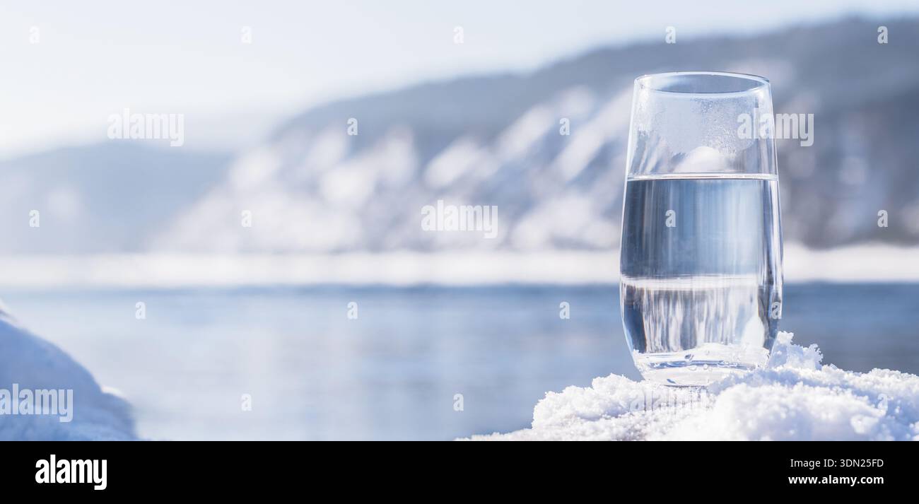 Un verre rempli d'eau propre repose sur un lit de neige. En arrière-plan, un paysage hivernal montre des collines et une rivière. La lumière du soleil brille sur le Banque D'Images