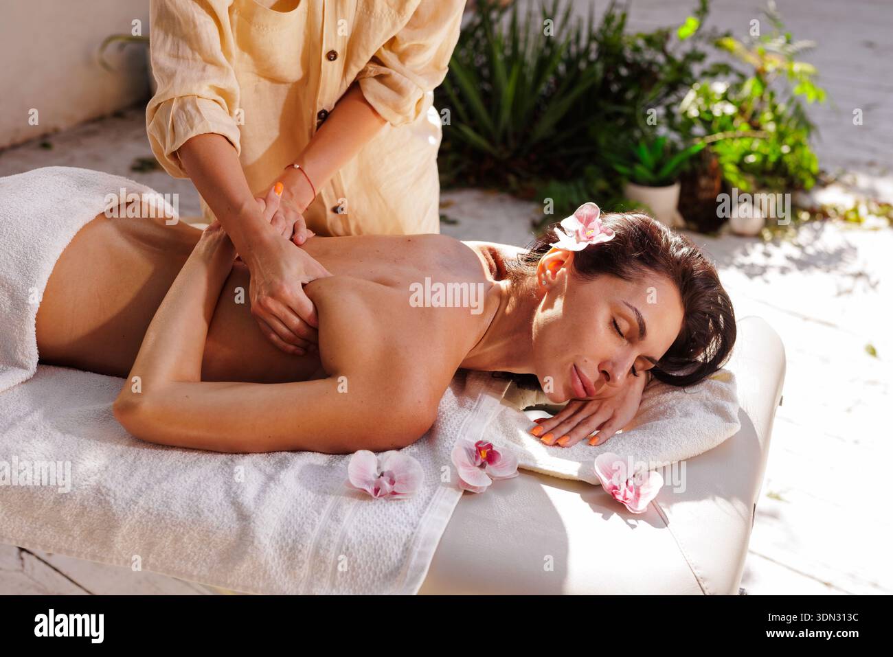 Femme recevant un massage professionnel du dos sur la table de spa à l'extérieur avec des fleurs d'orchidée et la lumière du soleil. Idéal pour la publicité bien-être, promotion Resort, an Banque D'Images