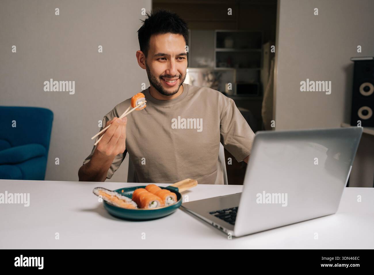 Joyeux jeune homme souriant profitant d'appels vidéo à distance ou de contenu en streaming sur ordinateur portable tout en prenant un repas de sushi, montrant le style de vie urbain moderne, travailler à partir de Banque D'Images