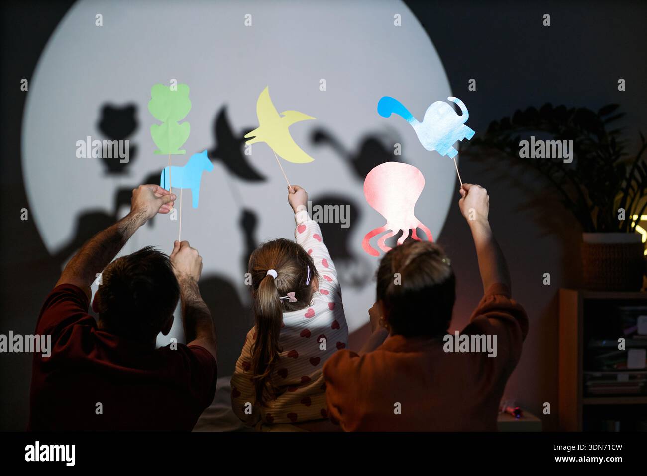 Homme caucasien, femme caucasienne et enfant caucasien tenant des marionnettes d'ombre en papier colorées devant un mur éclairé, créant des formes animales avec les mains levées ensemble Banque D'Images