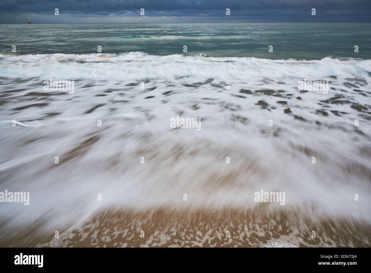 Paysage marin à longue exposition avec vagues mousseuses se lavant sur une plage de sable sous de sombres nuages orageux, un horizon calme et une atmosphère côtière sombre. Banque D'Images