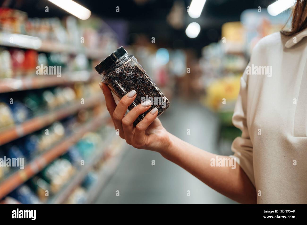 Femme shopper tient un pot rempli de feuilles de thé en vrac dans une allée d'épicerie, entouré d'emballages et de produits vibrants, mettant en évidence le Banque D'Images