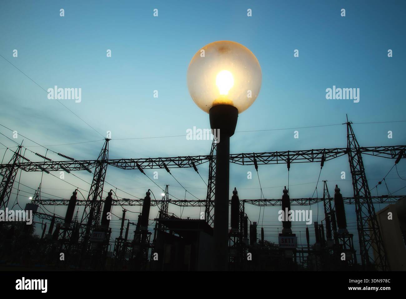 Un lampadaire lumineux se dresse au premier plan, silhouette contre un ciel crépusculaire. Derrière elle, une sous-station électrique complexe avec des tours et des fils i Banque D'Images