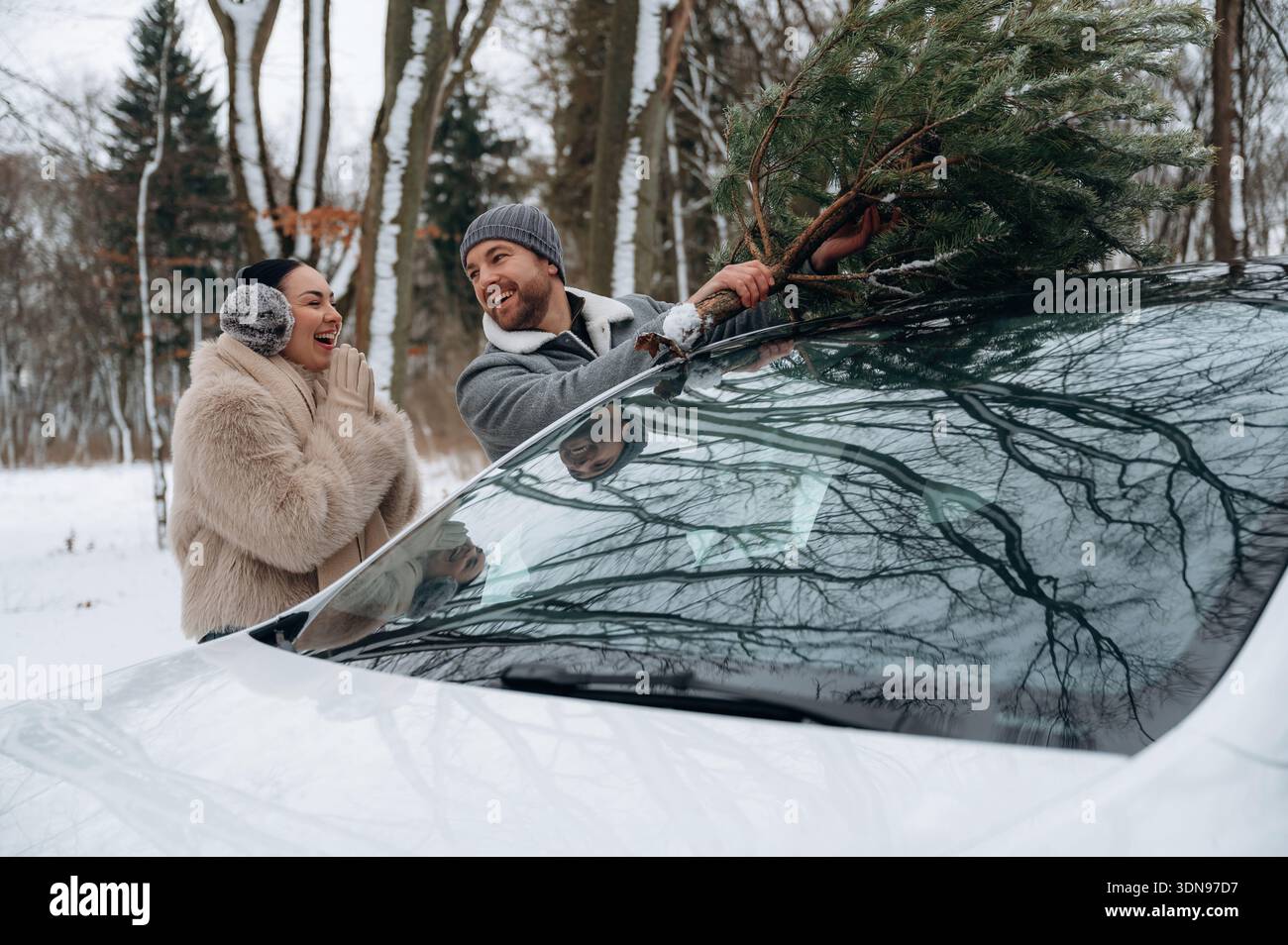 Heureux couple, habillé chaudement, est de sécuriser un arbre de Noël sur le toit de leur voiture lors d'une sortie hivernale, entouré par un paysage enneigé et grand t Banque D'Images
