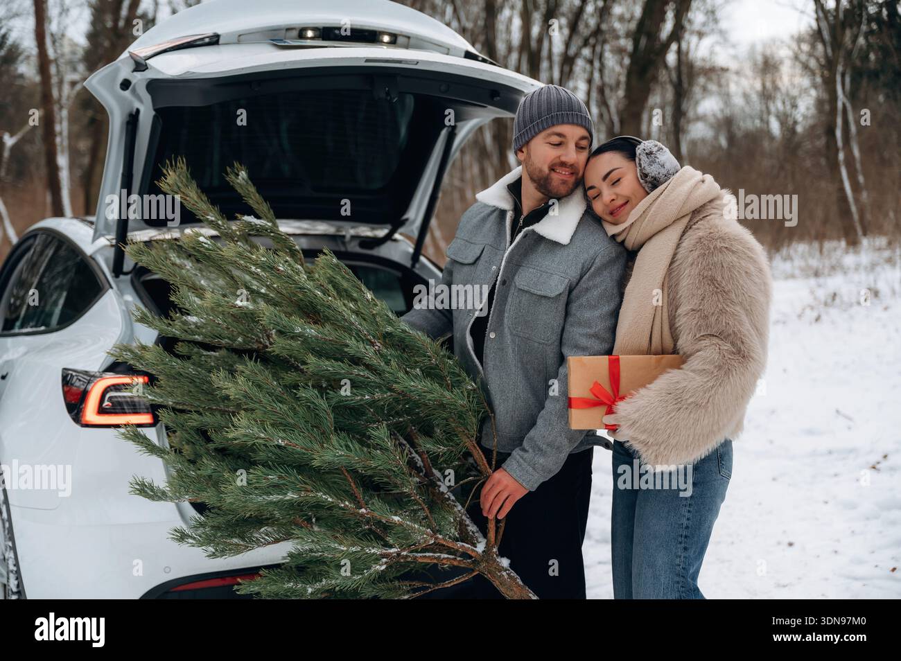 Femme asiatique et homme caucasien se préparent avec joie pour la saison des fêtes, chargeant un arbre de Noël et un cadeau dans le coffre de leur voiture, mis à nouveau Banque D'Images