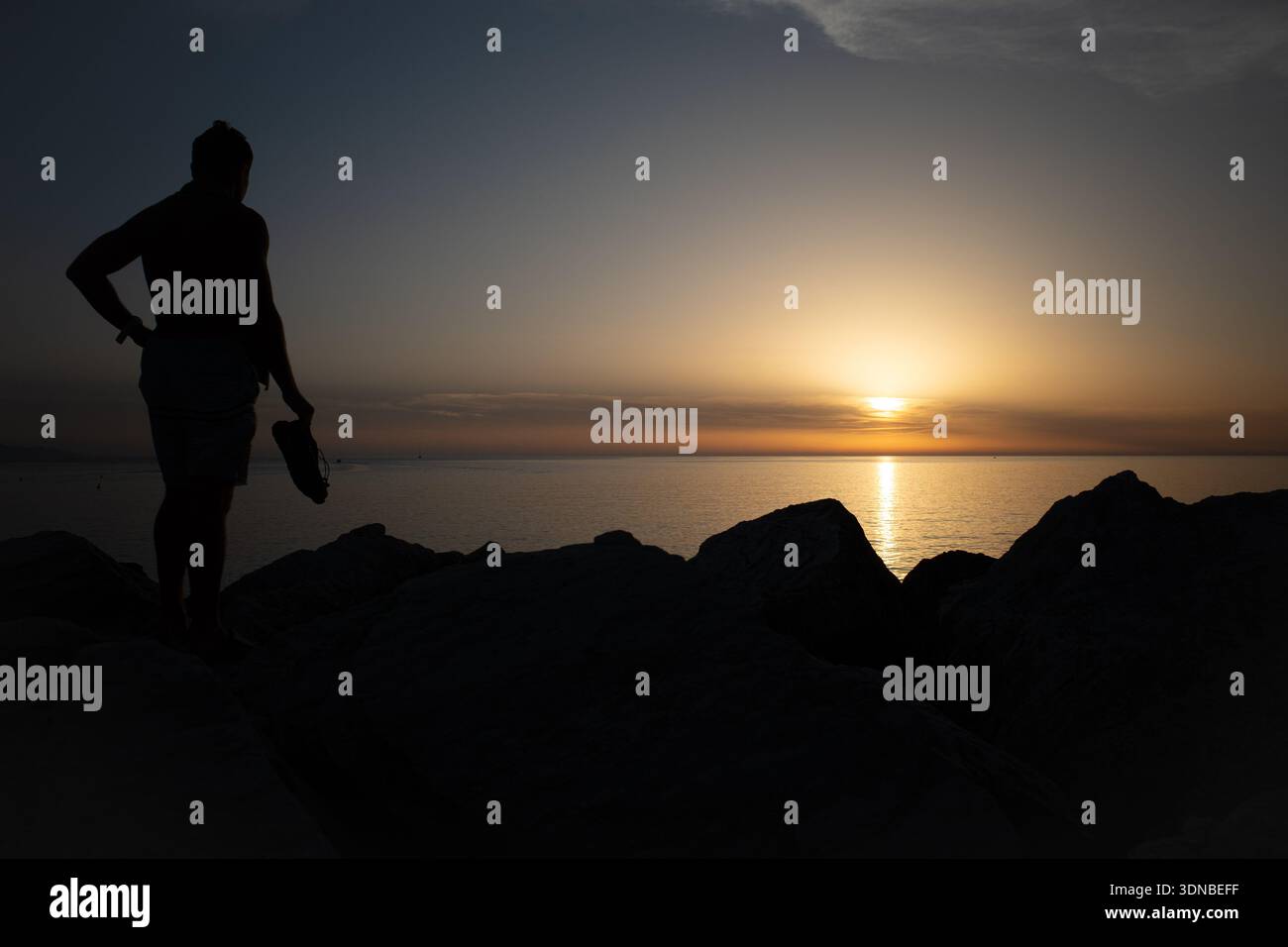 Silhouette de l'homme debout sur la rive rocheuse au coucher du soleil, tenant des chaussures et regardant le soleil doré se refléter sur l'horizon de la mer calme. Banque D'Images