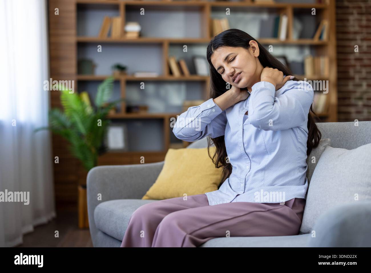 Jeune femme indienne assise sur un canapé massant le cou et les épaules, montrant des maux cervicaux et des tensions musculaires dues à une mauvaise posture, au stress et à la fatigue à la maison Banque D'Images