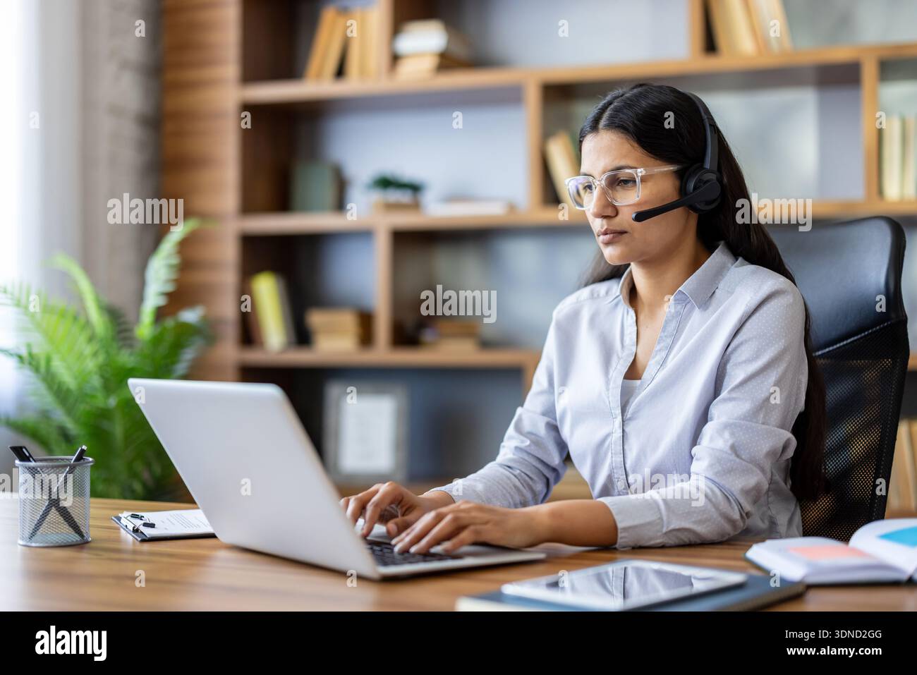 Jeune femme indienne fournissant un soutien à la clientèle ou une consultation en ligne, portant un casque avec un microphone, utilisant un ordinateur portable à un bureau à domicile avec une étagère en arrière-plan Banque D'Images