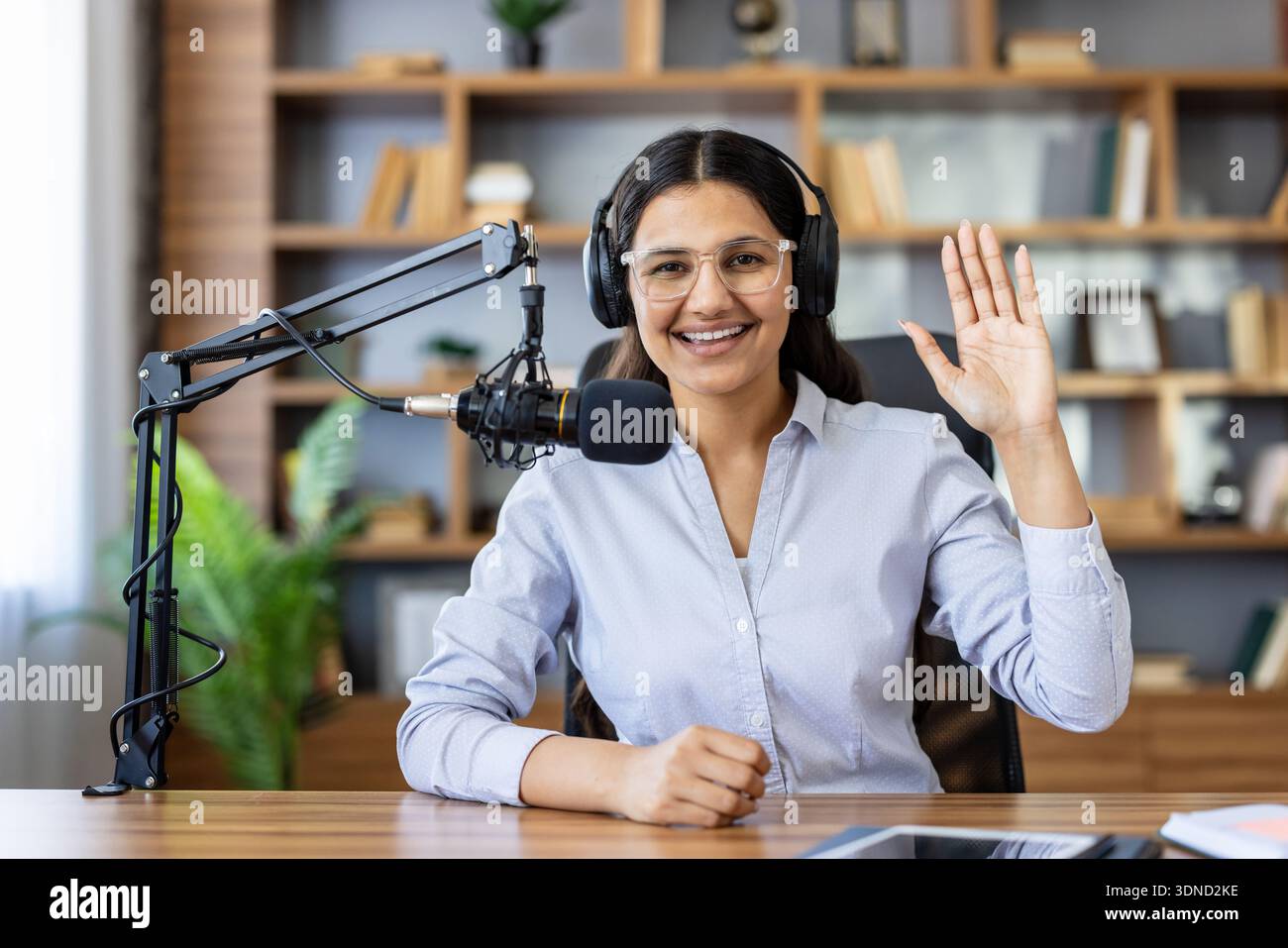 Jeune femme souriante portant des écouteurs et des lunettes enregistrant un podcast ou une diffusion en direct, assise à un bureau avec un microphone professionnel et agitant la main au public Banque D'Images