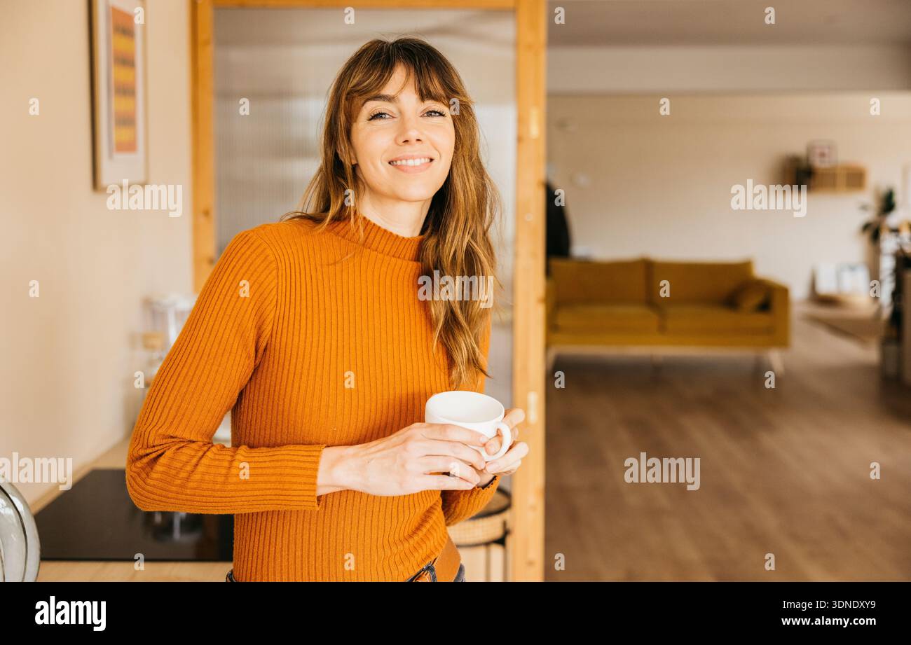 Femme souriant tout en tenant une boisson chaude, profitant d'une matinée domestique à la maison Banque D'Images