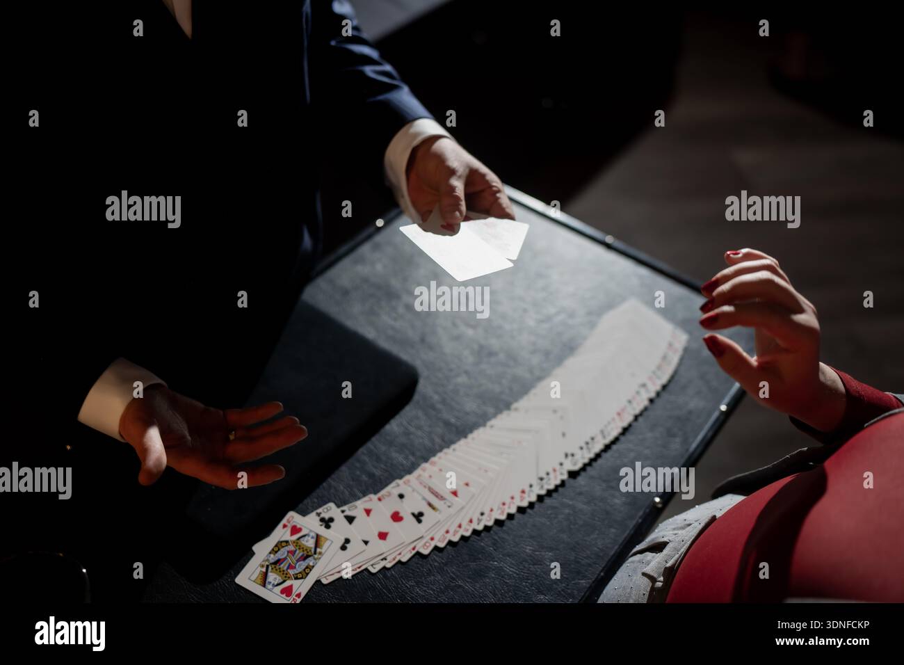 Magicien effectuant un tour de carte à table avec pont en éventail, tenant des cartes à jouer lumineuses tandis que la femme en robe rouge regarde sous les projecteurs dramatiques Banque D'Images
