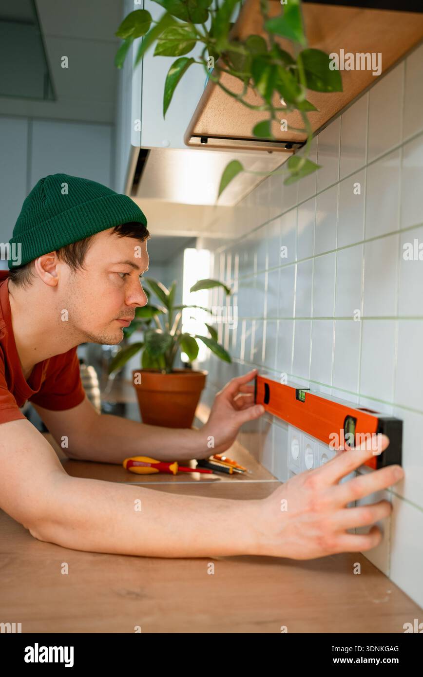 Homme utilisant un niveau à bulle inspectant l'installation de la prise électrique dans une cuisine moderne, assurant l'alignement horizontal pendant l'amélioration et la rénovation de la maison, en se concentrant sur les détails Banque D'Images