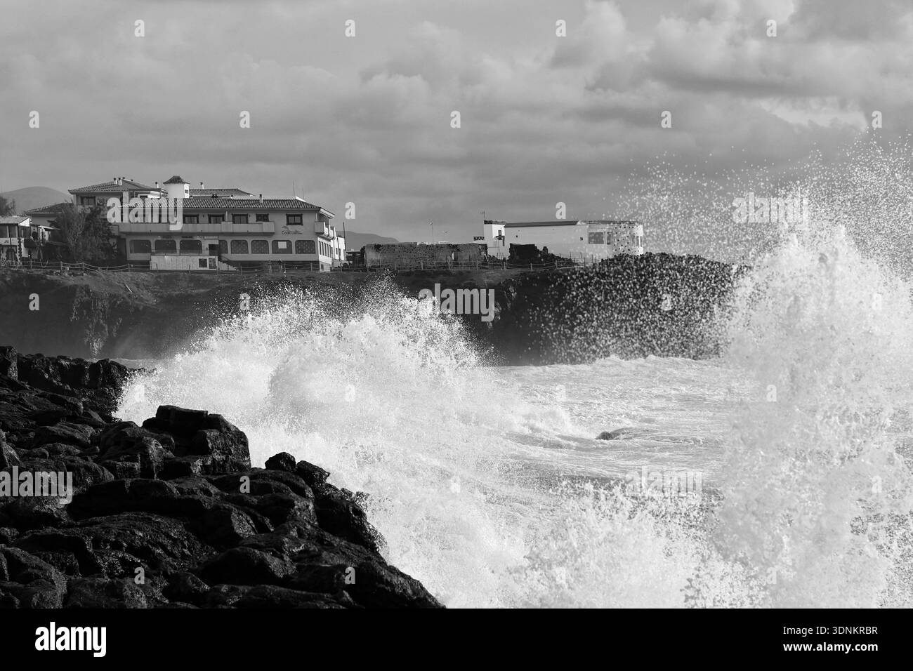 Photo en noir et blanc d'une mer orageuse avec de hautes vagues saccadées, des rochers et des maisons en arrière-plan Banque D'Images
