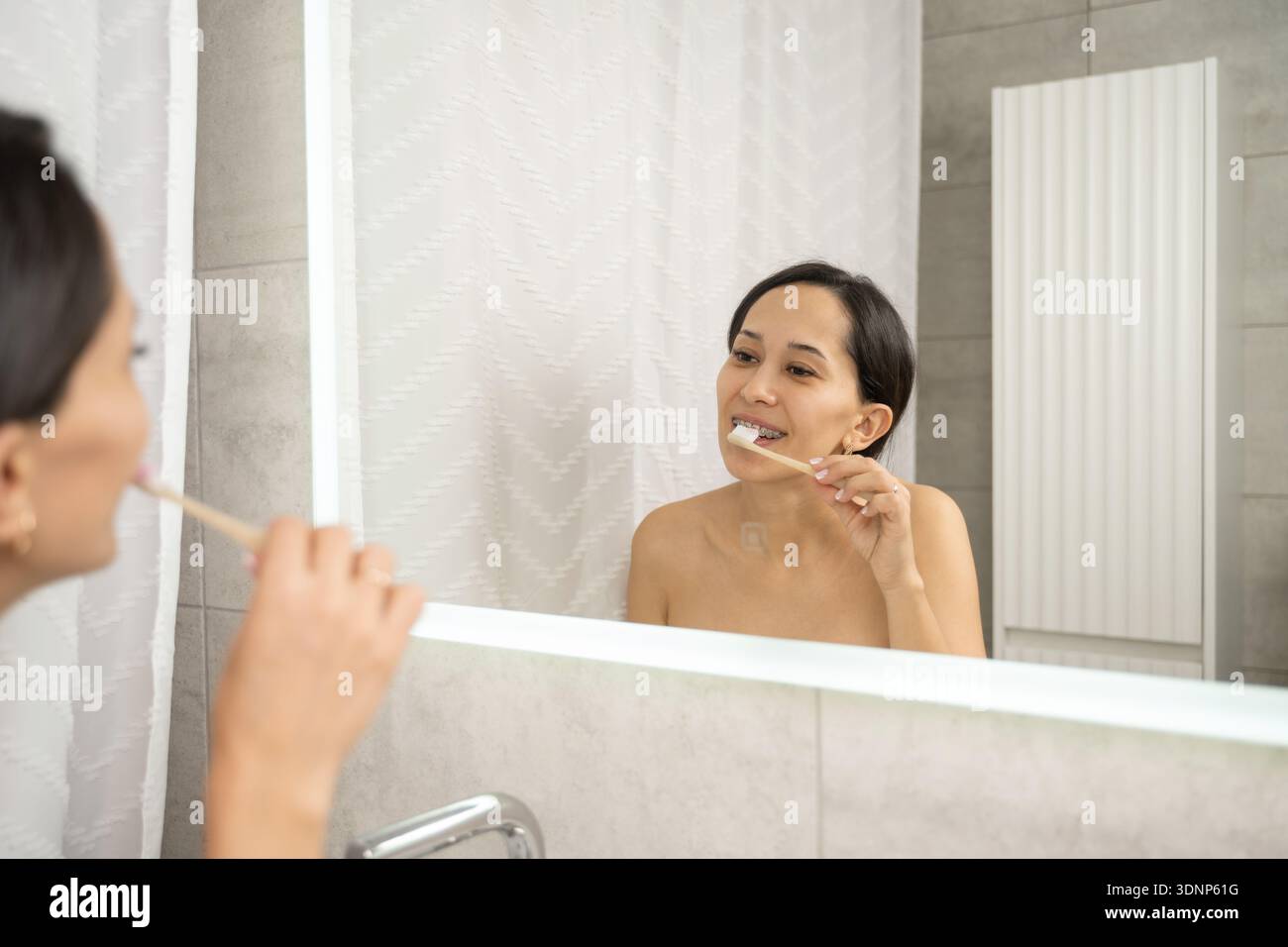 Une femme se tient devant un miroir de salle de bain se brossant les dents, pratiquant une bonne hygiène bucco-dentaire. Son reflet est visible dans le miroir, montrant un clea Banque D'Images