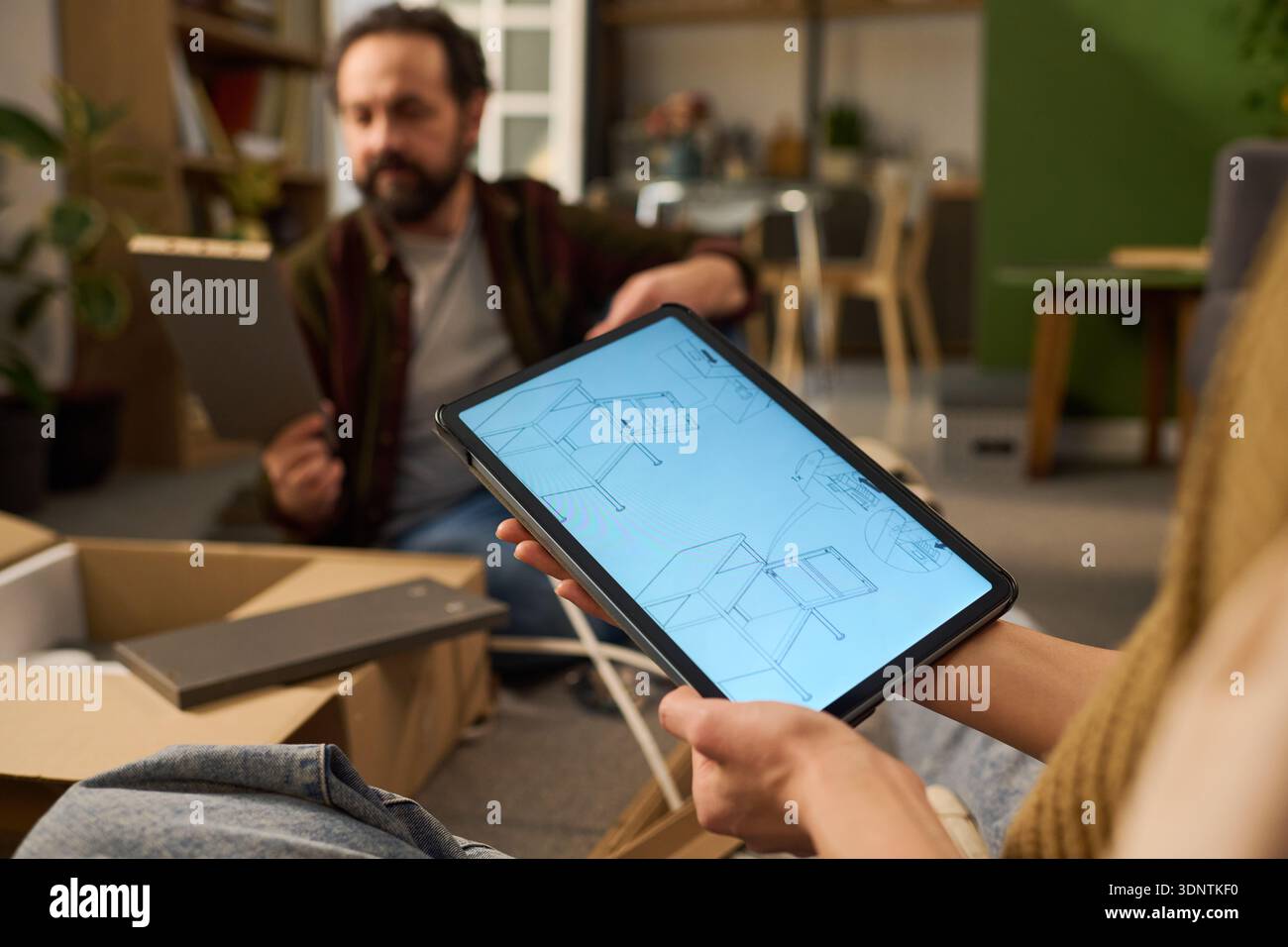 Femme jeune adulte caucasienne tenant une tablette numérique affichant des instructions d'assemblage de meubles tandis que l'homme d'âge moyen caucasien assemblant des meubles dans un cadre de maison moderne Banque D'Images