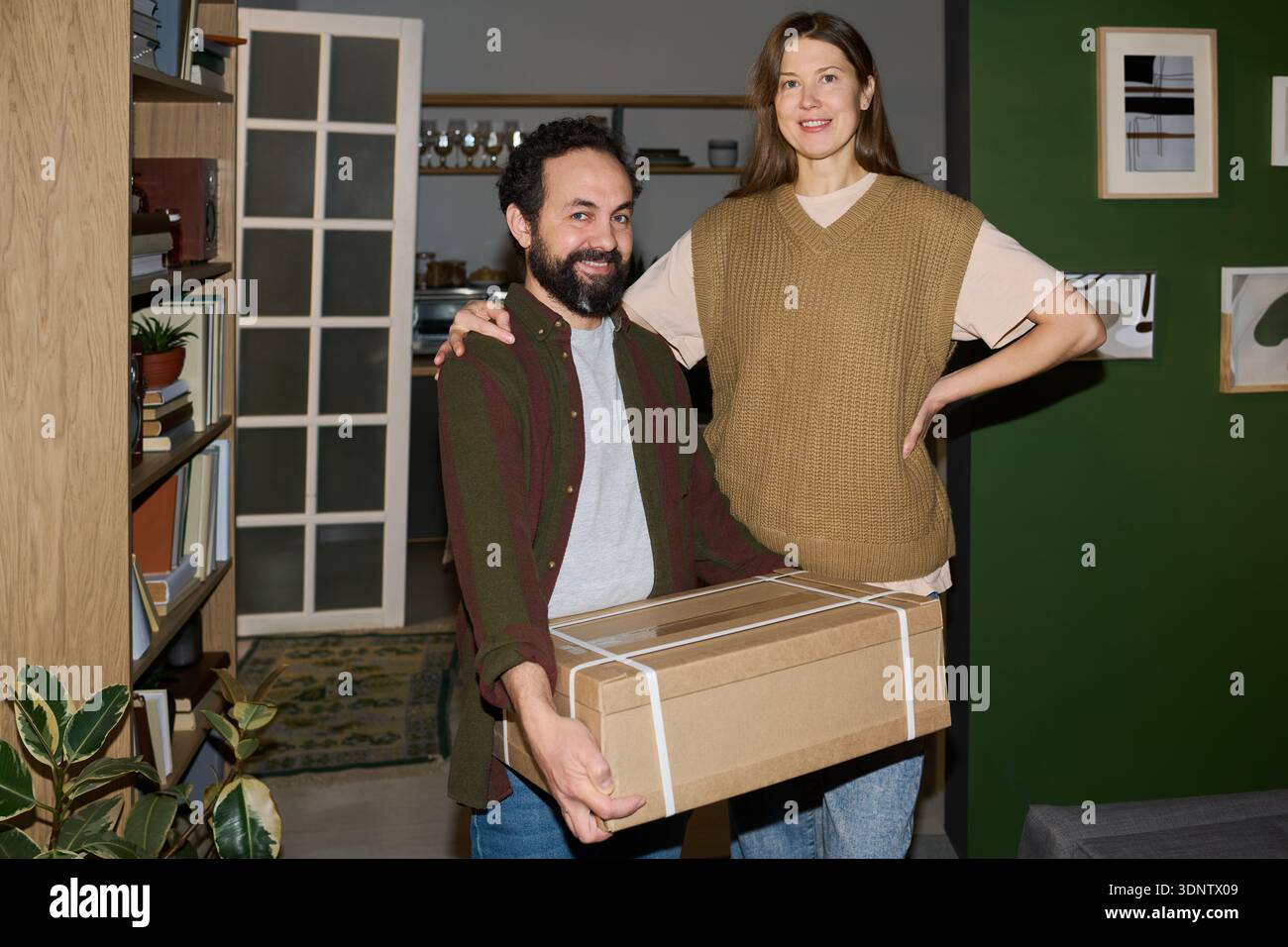 Portrait de jeune homme adulte caucasien souriant tout en tenant une boîte en carton, debout à côté de jeune femme adulte caucasienne avec la main sur la hanche Banque D'Images