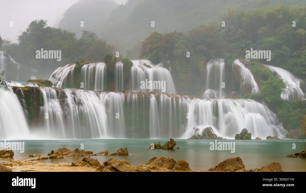 La cascade de Ban Gioc coule dans de larges rideaux étagés sur des falaises calcaires dans une piscine turquoise calme, avec des rochers et des bancs de sable au premier plan. Banque D'Images