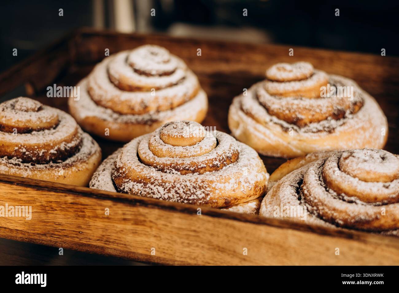 Délicieux petits pains à la cannelle disposés dans un plateau en bois rustique, saupoudrés de sucre en poudre. Cette vitrine alléchante de pâtisseries sucrées est prête à tenter le café Banque D'Images