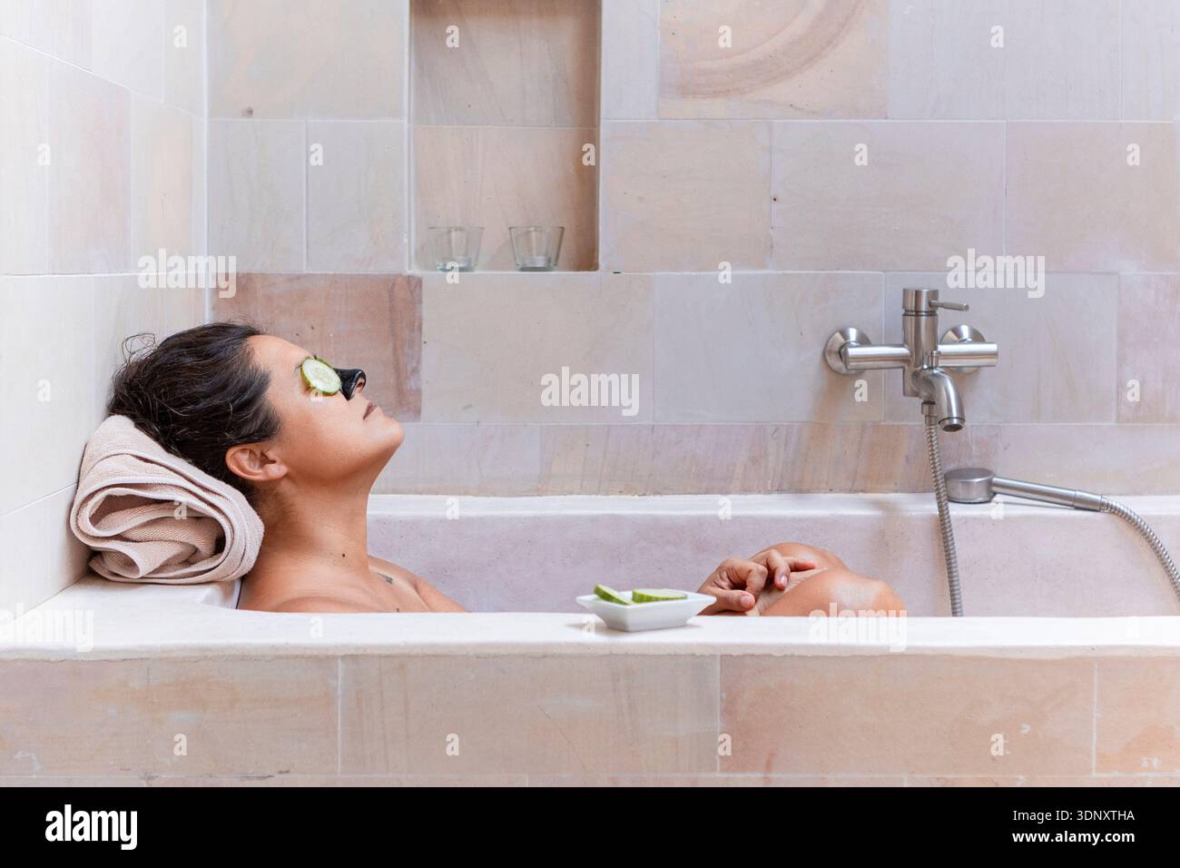 Une femme se détend dans une baignoire contemporaine, profitant d'un moment de sérénité avec des tranches de concombre sur ses yeux. Le cadre reflète un accent mis sur les soins personnels et Banque D'Images