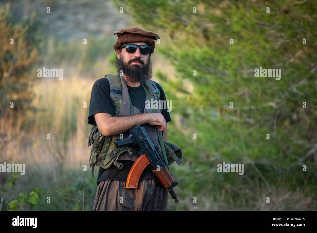 Un homme est vêtu d'un équipement de style militaire, avec gilet tactique et lunettes de soleil, tenant un fusil airsoft tout en se tenant debout dans un set naturel d'extérieur Banque D'Images