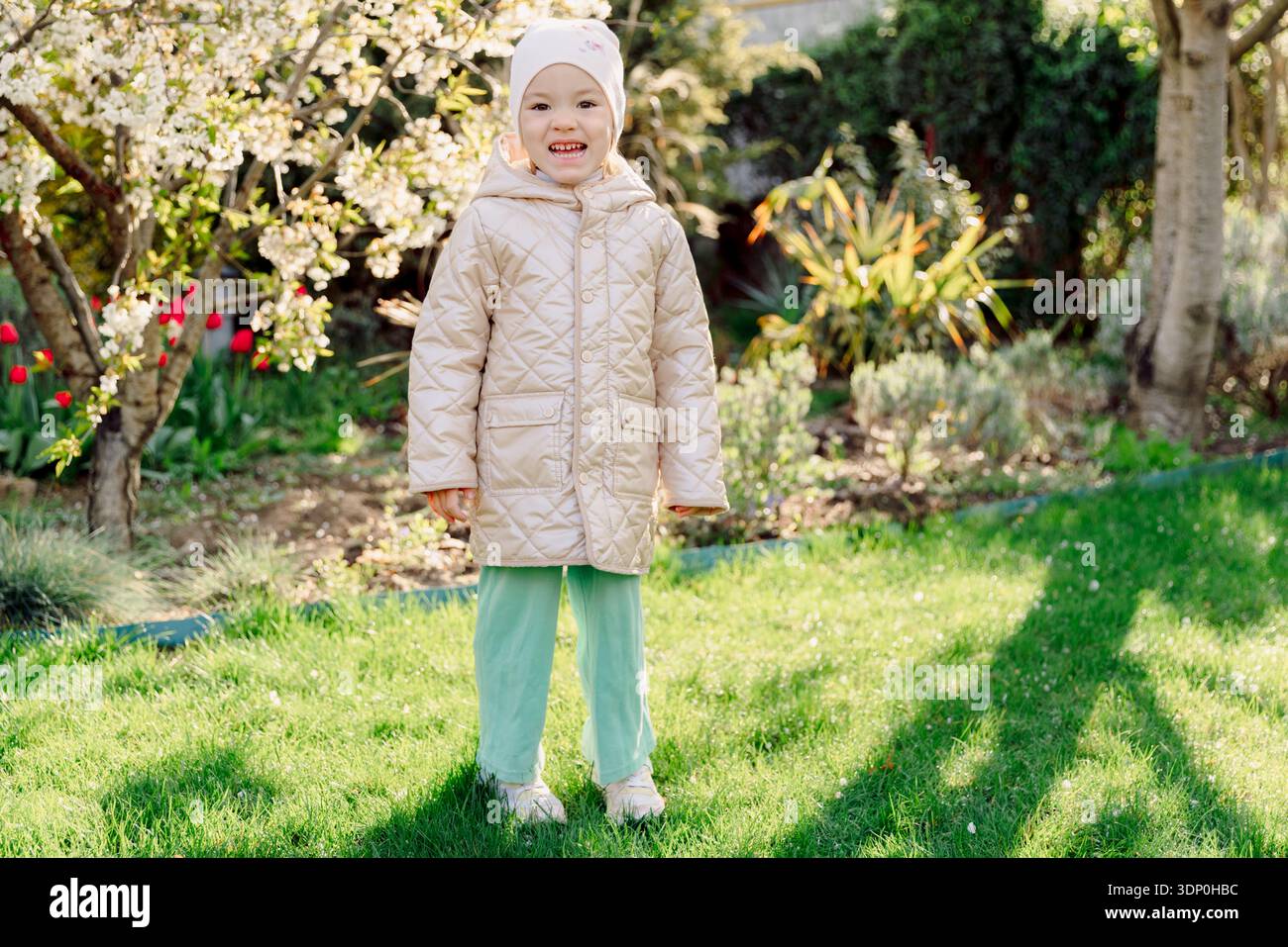 Tout-petit souriant debout dans un jardin printanier fleuri, la lumière du soleil et la verdure vibrante créant un moment de joie en famille. Banque D'Images