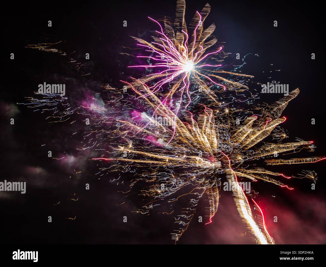Explosion de feu d'artifice doré en bas à droite, éclatement coloré plus petit avec noyau blanc brillant au-dessus, lueur rouge s'estompant à droite, nuit noire profonde percée par la lumière. Banque D'Images