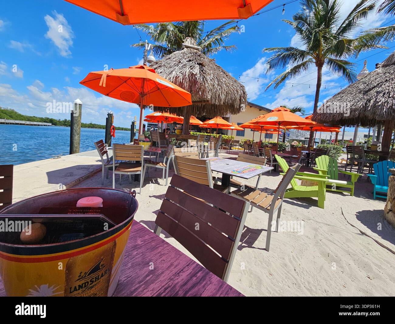 USAGE ÉDITORIAL EXCLUSIF - MARGARITAVILLE HOLLYWOOD BEACH, FIVE O'CLOCK SOMEWHERE BAR AND GRILL : salle à manger extérieure animée dans un bar de plage tropical en Floride, avec parasols orange et cabanes tiki au toit de chaume. Des chaises Adirondack colorées et des tables en bois sont disposées sur du sable blanc le long d'une voie navigable sous un ciel bleu clair. - Image de stock capturée avec un smartphone