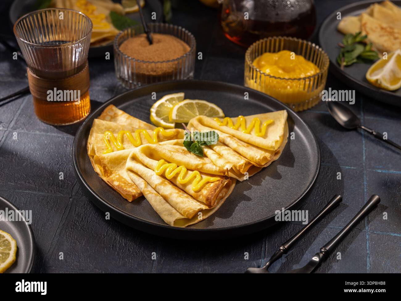 Fines crêpes pliées sur une assiette noire surmontée de caillé de citron brillant, de menthe fraîche et d’une tranche de citron, servies avec du sucre, du thé chaud et du caillé supplémentaire pour le brunch Banque D'Images