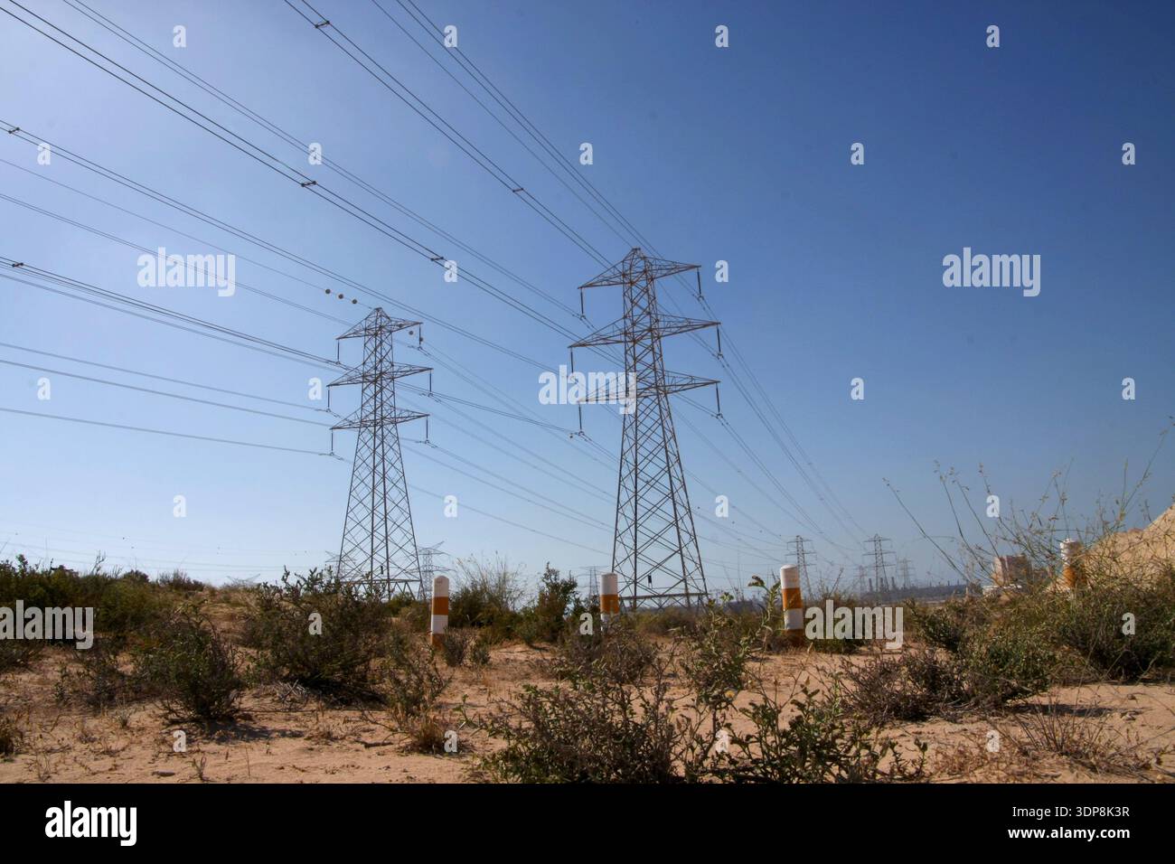 Pylônes de transmission haute tension et lignes électriques traversant un paysage désertique sec et sablonneux avec des arbustes verts sous un ciel dégagé. Banque D'Images