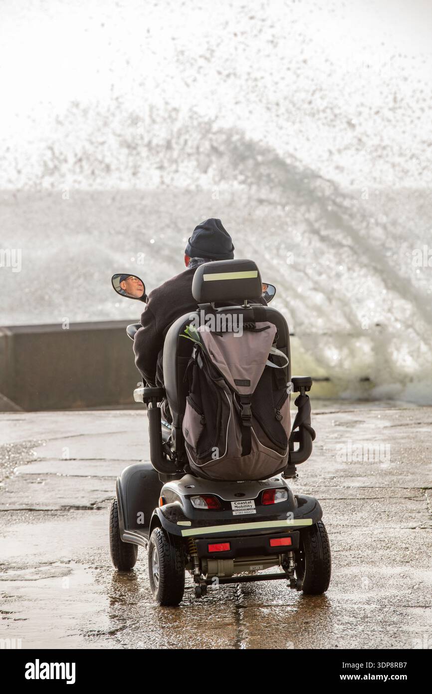 Homme handicapé âgé en fauteuil roulant motorisé assis au bord de la mer regardant les vagues se briser sur les défenses maritimes pendant une tempête hivernale. Banque D'Images
