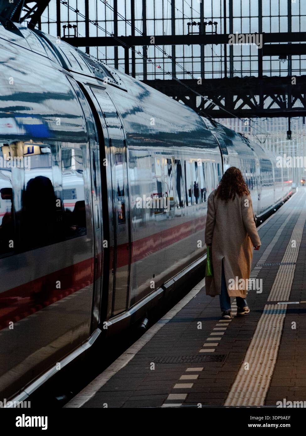 Amsterdam, pays-Bas - 1er mars 2025 : femme marchant sur le quai à côté d'un train moderne élégant, reflets métalliques sur sa surface, sous le toit d'une gare Banque D'Images