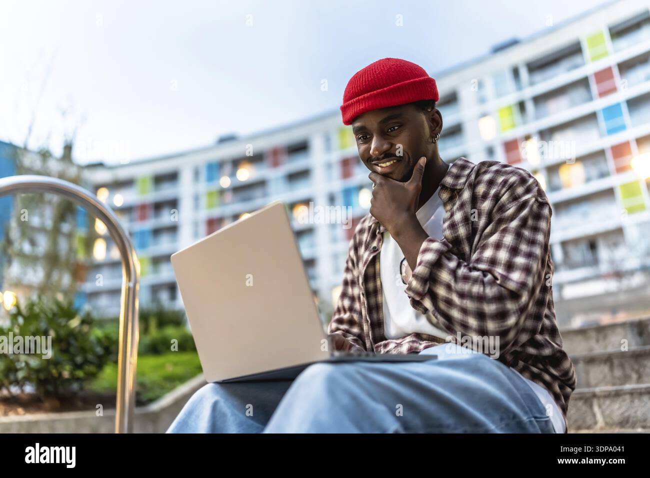 Jeune homme noir assis sur les marches de l'appartement avec un ordinateur portable, souriant et réfléchi comme il travaille ou étudie à l'extérieur, incarnant le travail à distance urbain moderne et Banque D'Images