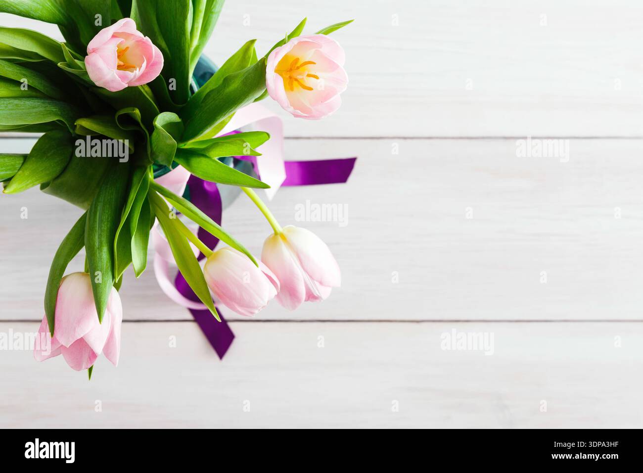 Tulipes roses dans un vase avec feuilles vertes et ruban violet sur surface en bois Banque D'Images