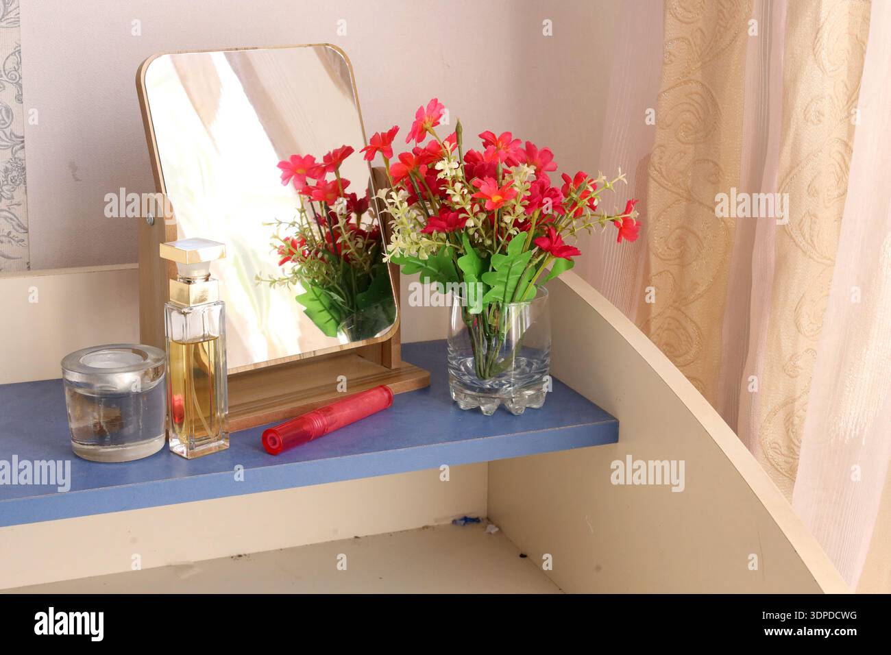 Disposition de table de toilette avec un miroir de bureau en bois reflétant des fleurs, une bouteille de parfum, une bougie et un rouge à lèvres rouge sur une surface bleue. Banque D'Images