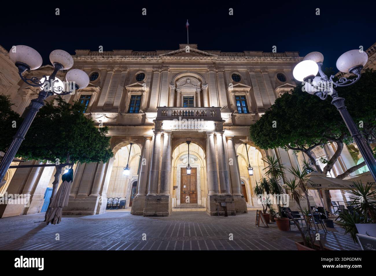 Façade néoclassique illuminée de la Bibliothèque nationale de Malte à la Valette, avec entrée voûtée, colonnes, lampadaires et arbres de la cour la nuit Banque D'Images