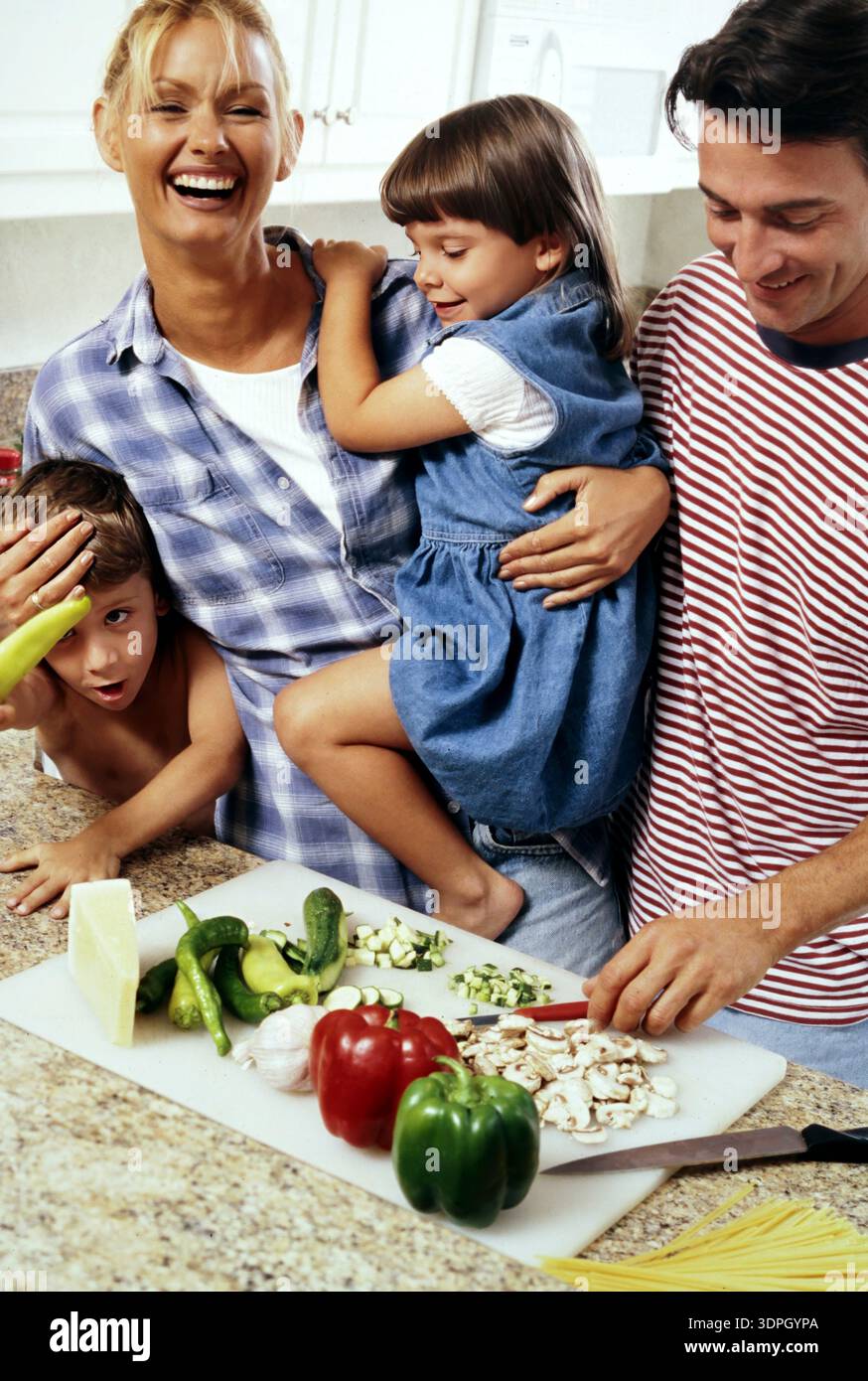 Une famille s'amuse dans la cuisine tout en préparant un repas. Deux enfants s'impliquent, dont un assis sur le dos d'un parent. Légumes frais Banque D'Images