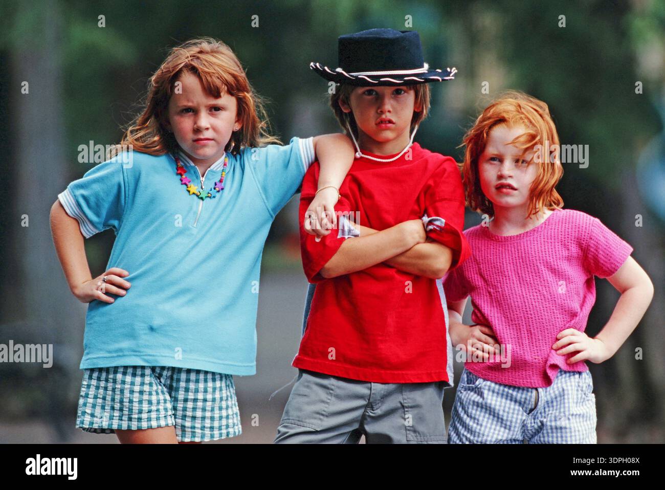 Trois enfants se tiennent côte à côte dehors. Deux filles aux cheveux longs portent des vêtements décontractés tandis qu'un garçon au milieu a une expression sérieuse. Ils montrent Banque D'Images