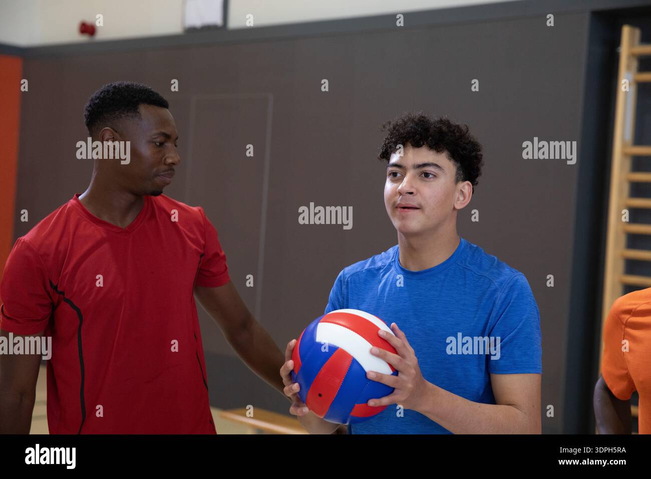 Divers coéquipiers masculins entraîneur d'adolescent tenant du volley tricolore dans la salle de gym, portant des chemises rouge-bleu Banque D'Images