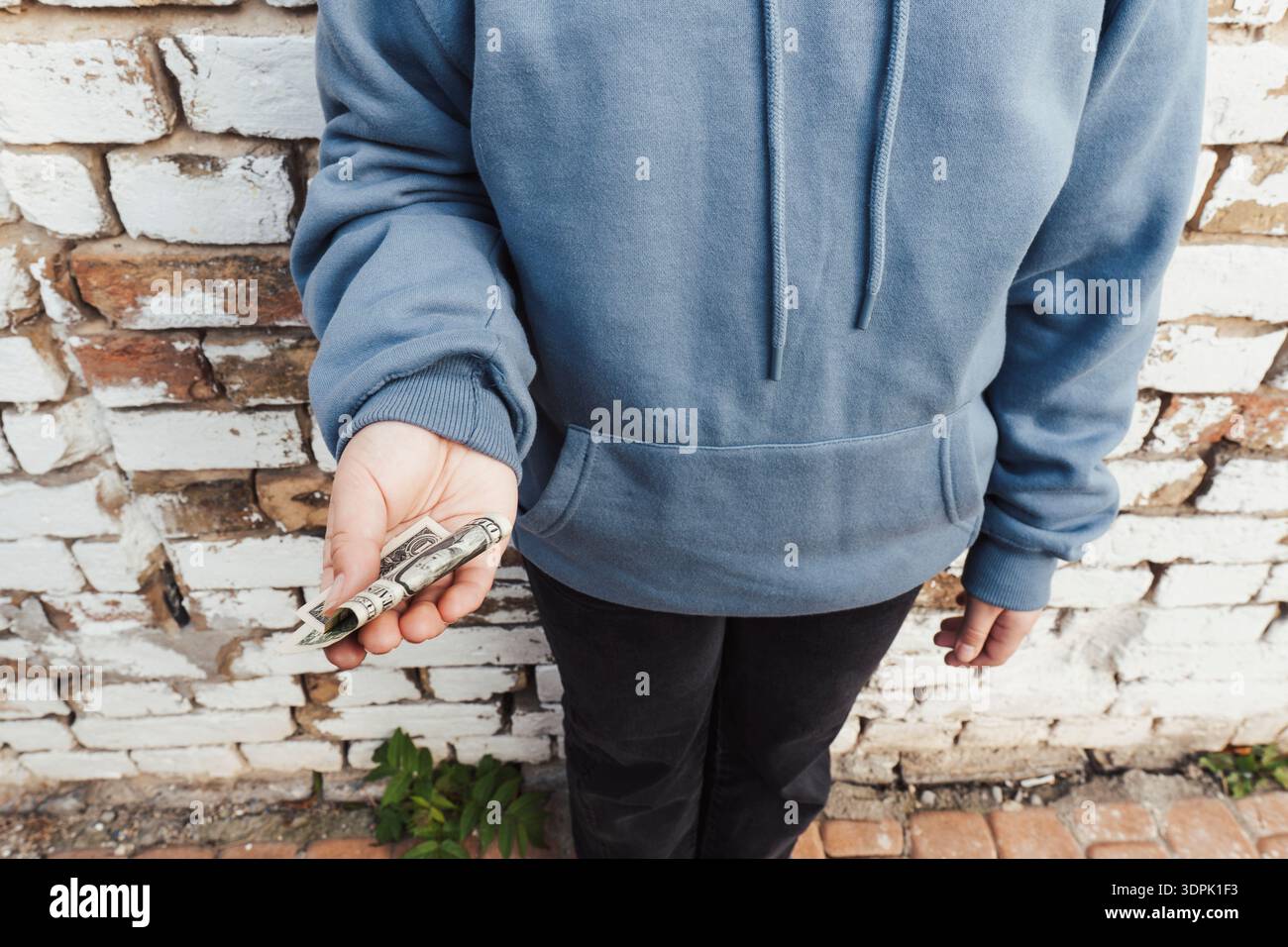 Gros plan d'une pauvre fille portant un sweat à capuche bleu tenant un billet d'un dollar plié. Notion d'inégalité sociale, de pauvreté et d'aide financière aux nécessiteux Banque D'Images