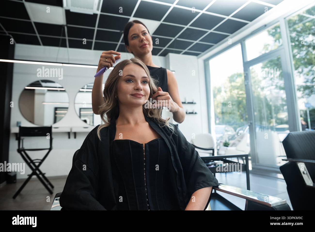 Une femme est assise dans une chaise de salon tandis qu'un styliste coiffe habilement ses cheveux. L'espace lumineux dispose de miroirs et d'un design épuré, améliorant le salon ex Banque D'Images