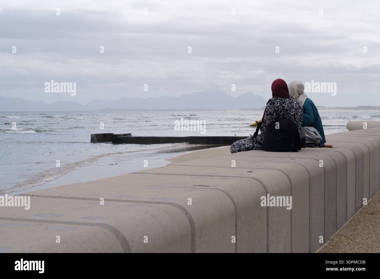 Deux amies anonymes assises sur un rebord passant du temps ensemble et partageant un moment à la plage de Strand, Cape Town, Afrique du Sud. Banque D'Images