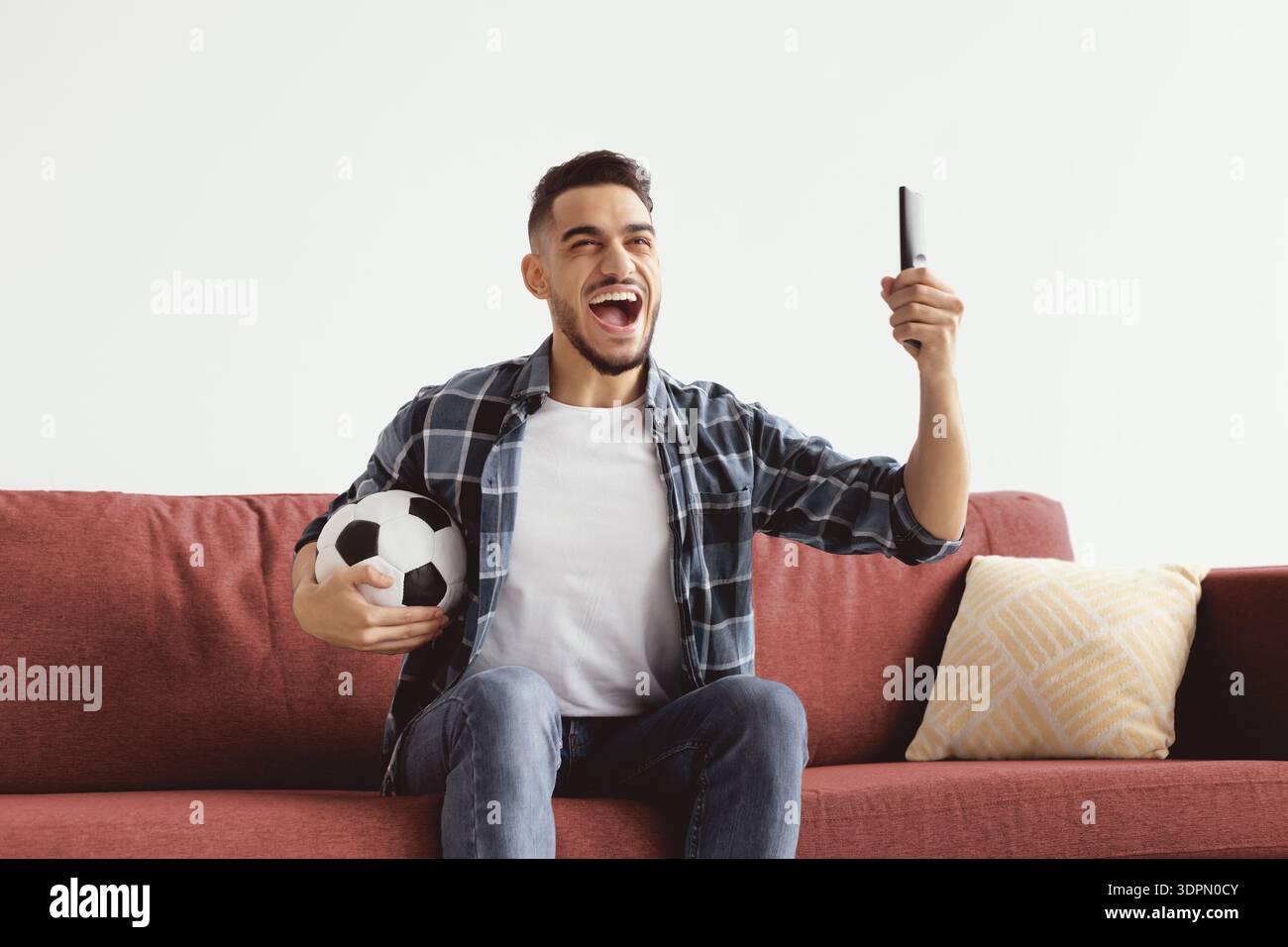 Man on Couch célèbre tout en tenant un ballon de football et un téléphone pendant un événement sportif Banque D'Images