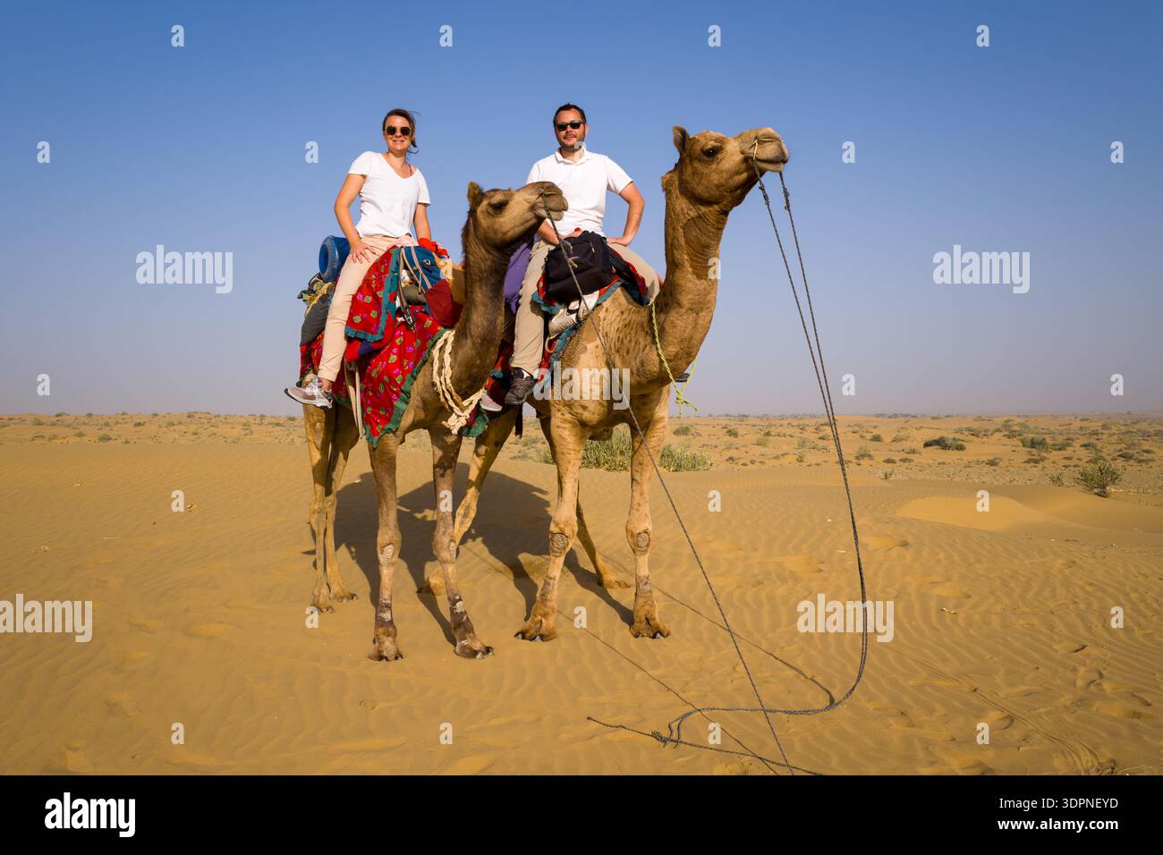 Jaisalmer, Inde - 29 mars 2022 : deux personnes montent à dos de chameaux avec des selles colorées dans le paysage sablonneux du désert du Thar sous un ciel dégagé. Banque D'Images